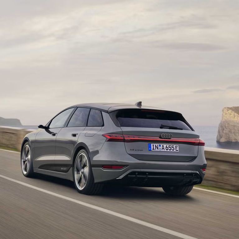 Schräge Heckaufnahme des A6 Avant e-tron, welcher auf einer Straße entlang des Meeres fährt. Im Hintergrund sind Felsen und Berge zu erkennen. 