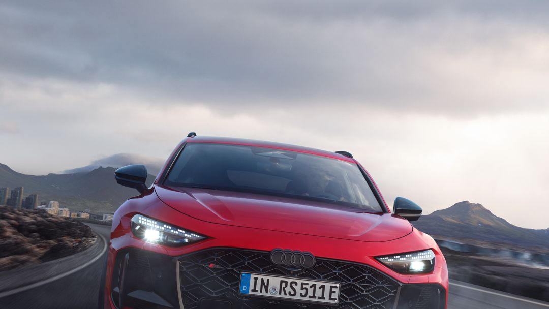 Ein roter Audi RS 5 Avant fährt auf einer Straße in einer bergigen Landschaft. Der Himmel ist bewölkt.