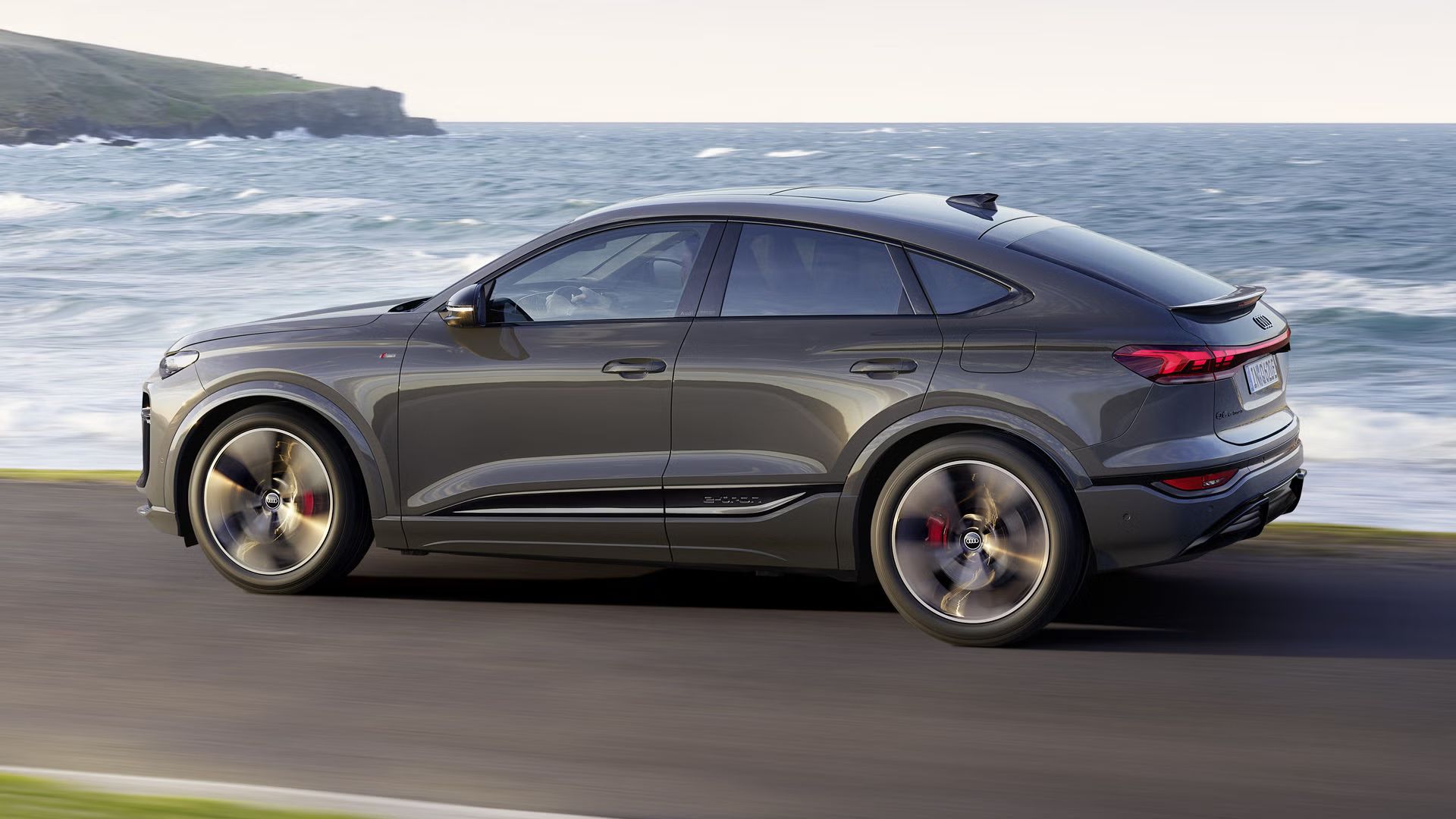 Seitliche Aufnahme des Q6 Sportback e-tron, welcher auf einer Straße entlang des Meeres fährt.