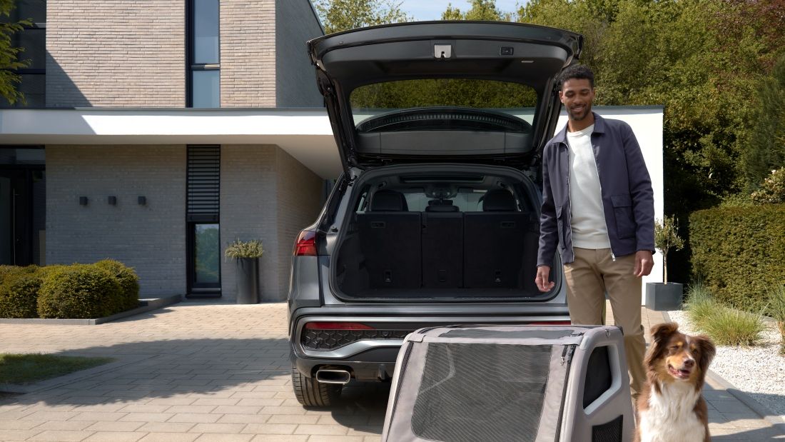 Sitzender Australian Shepherd neben der Audi Hundebox neben seinem lächelnden Herrchen vor dem geöffneten Kofferraum eines Audi Q5