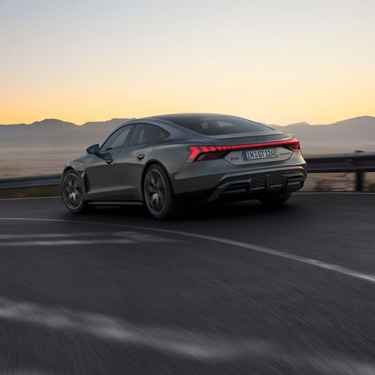 Ein grauer Audi RS e-tron GT fährt auf einer kurvigen Bergstraße bei Sonnenuntergang, mit leuchtenden Rücklichtern.