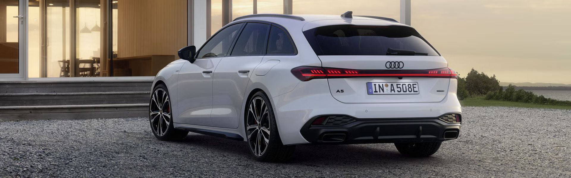 Der A5 Avant e-hybrid steht in einer schrägen Heckperspektive auf einer Kiesfläche vor einem modernen Wohnhaus.