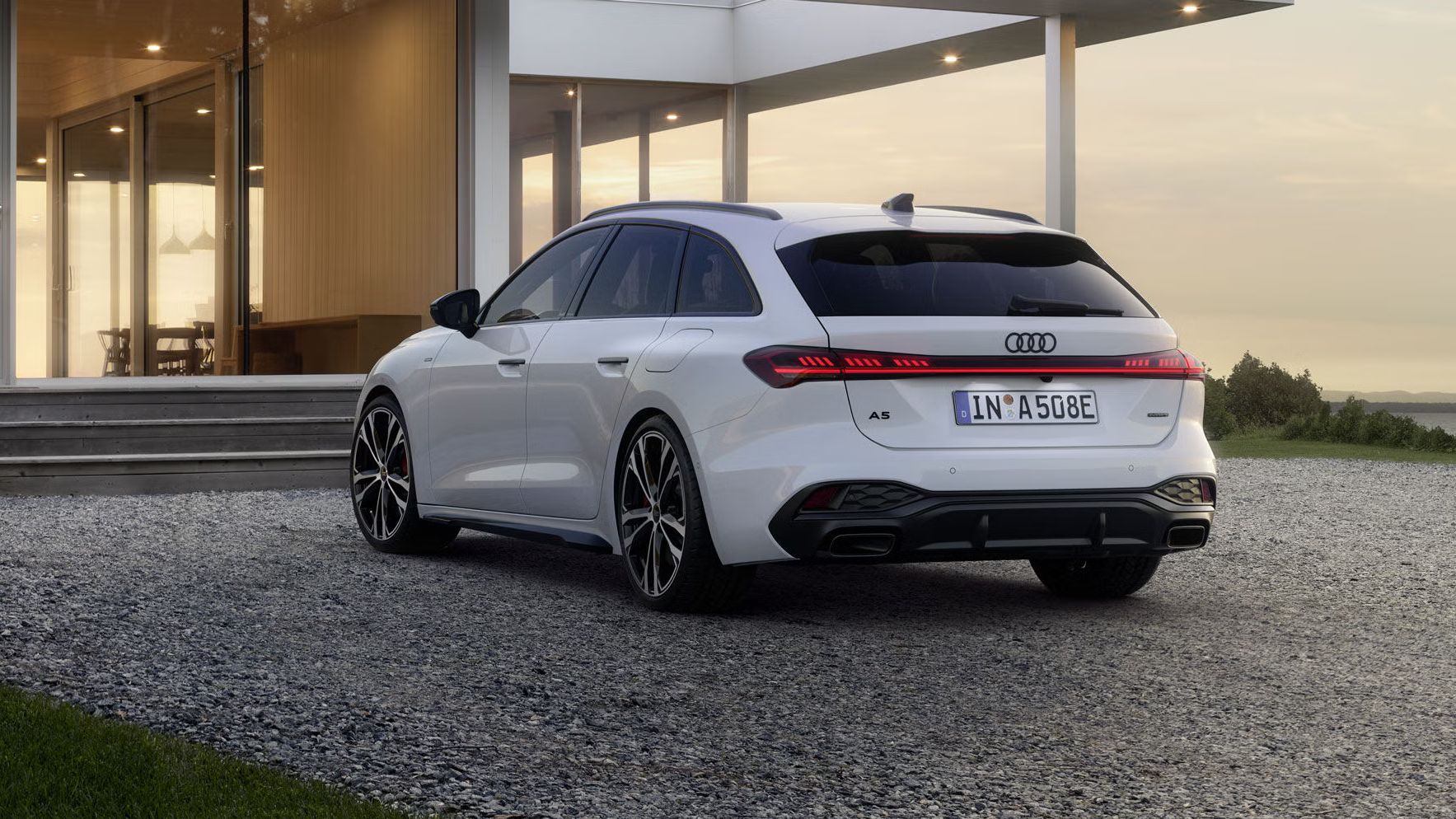 Der A5 Avant e-hybrid steht in einer schrägen Heckperspektive auf einer Kiesfläche vor einem modernen Wohnhaus.
