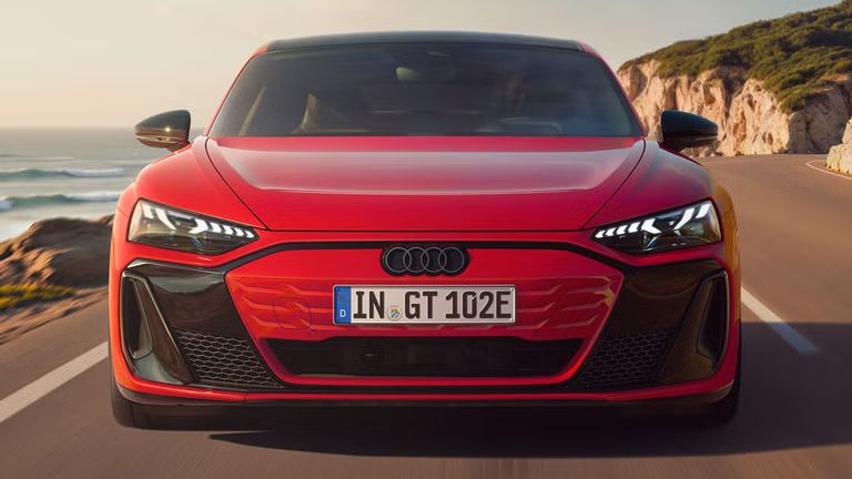 Frontansicht eines roten Audi e-tron GT quattro, der auf einer Küstenstraße fährt. Im Hintergrund sind Klippen, das Meer und der Himmel zu erkennen. 