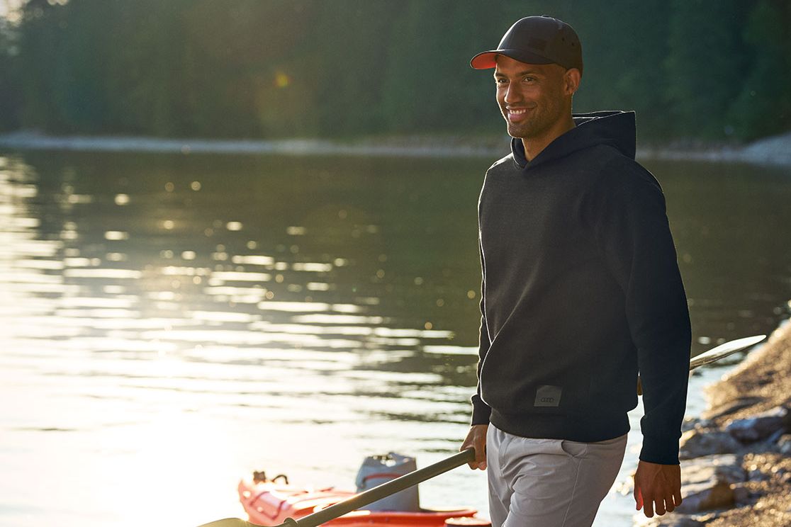 Eine Person in Sportkleidung steht am steinigen Ufer eines Sees und hält ein Paddel in der Hand. Neben ihr liegt ein orangefarbenes Kajak halb im Wasser, während die Sonne tief steht und auf der Wasseroberfläche glitzert. Im Hintergrund ist ein dichter Wald am anderen Seeufer zu sehen.