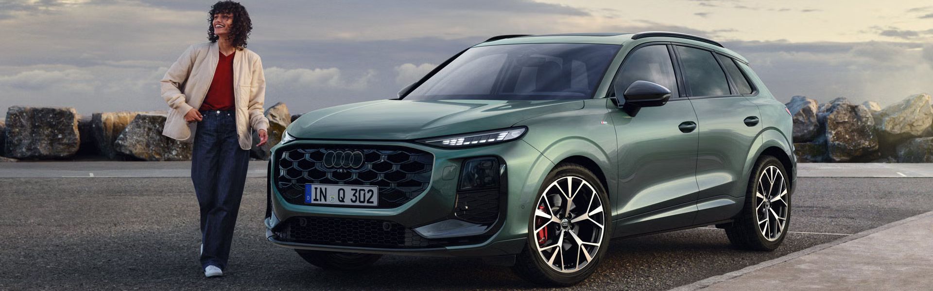 Seitliche, vordere Ansicht auf den geparkten Q3 SUV an einer Küstenstraße, daneben eine junge Frau. 