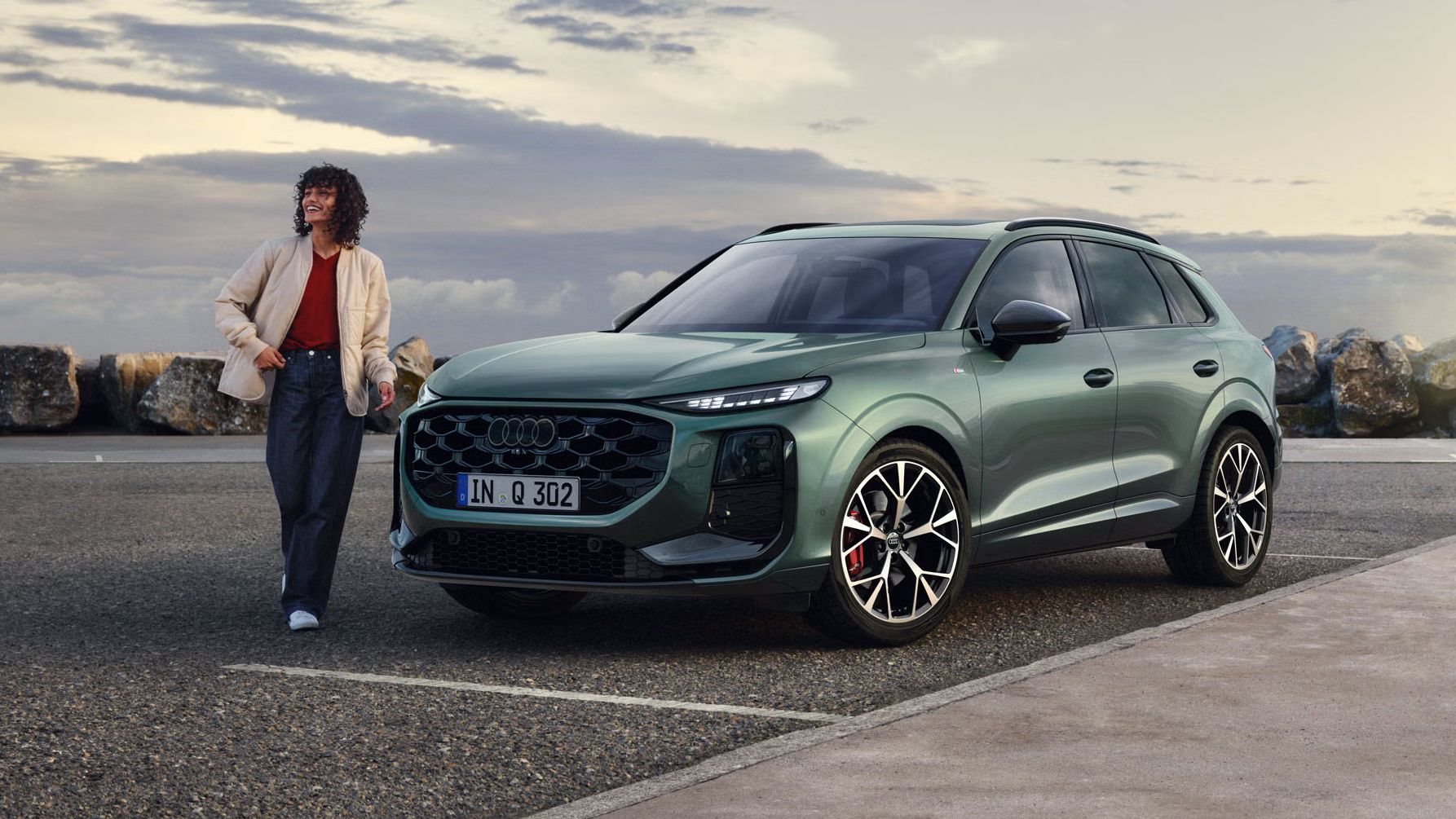 Seitliche, vordere Ansicht auf den geparkten Q3 SUV an einer Küstenstraße, daneben eine junge Frau. 