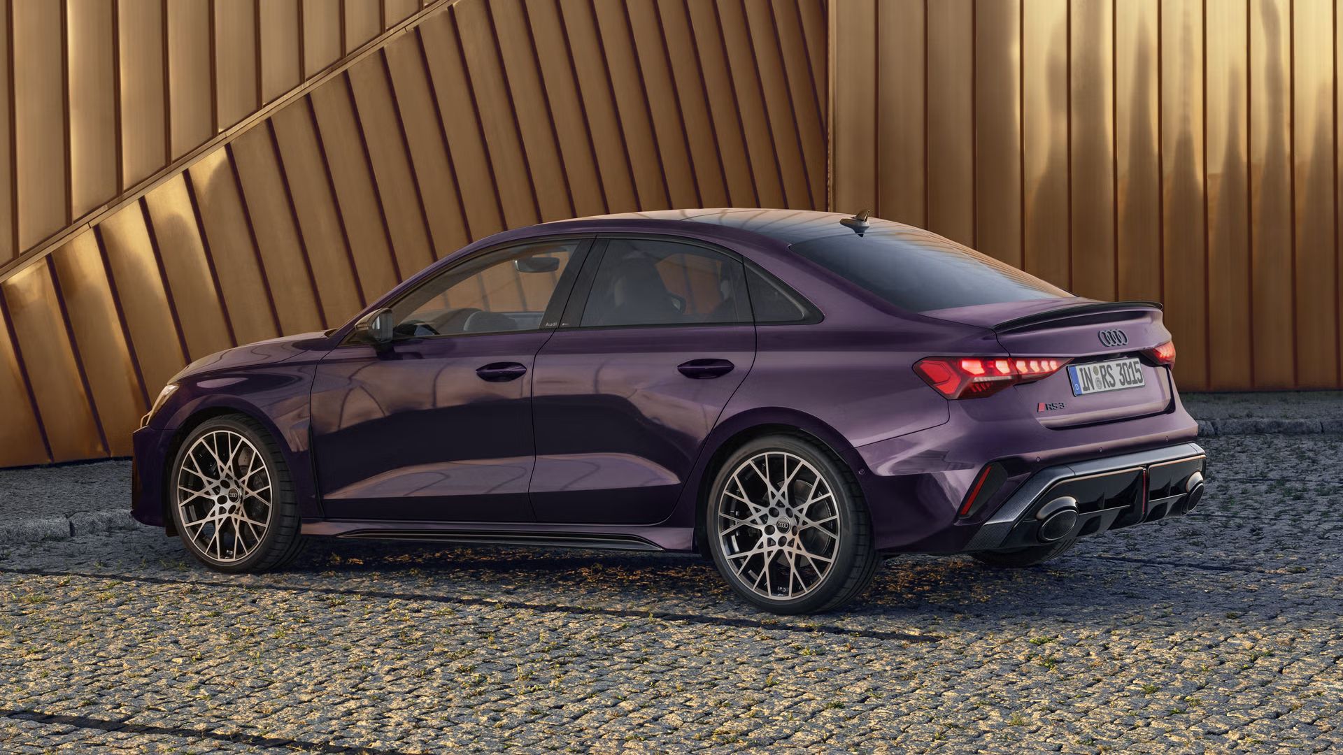 Die RS 3 Limousine mit Indivuduallackierung in Merlin Perleffekt von der Seite parkend auf Kopfsteinpflaster, im Hintergrund Umrisse eines modernen Gebäudes.