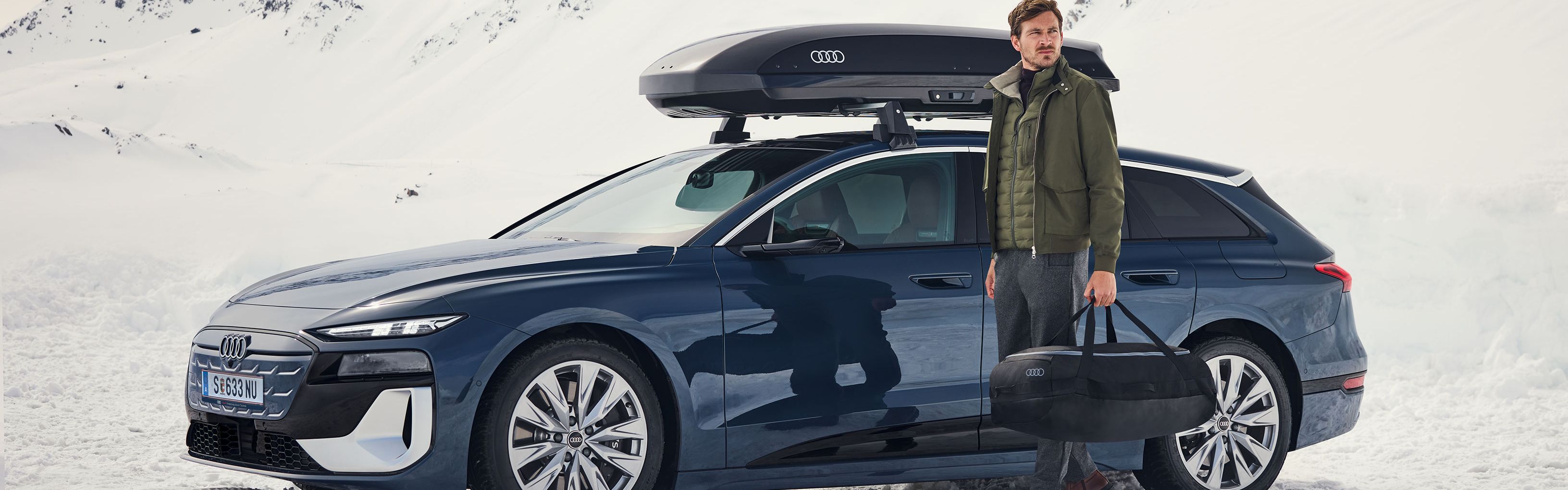 Ein Audi S6 mit Dachbox parkt in einer winterlichen Berglandschaft, ein Mann mit Tasche steht davor