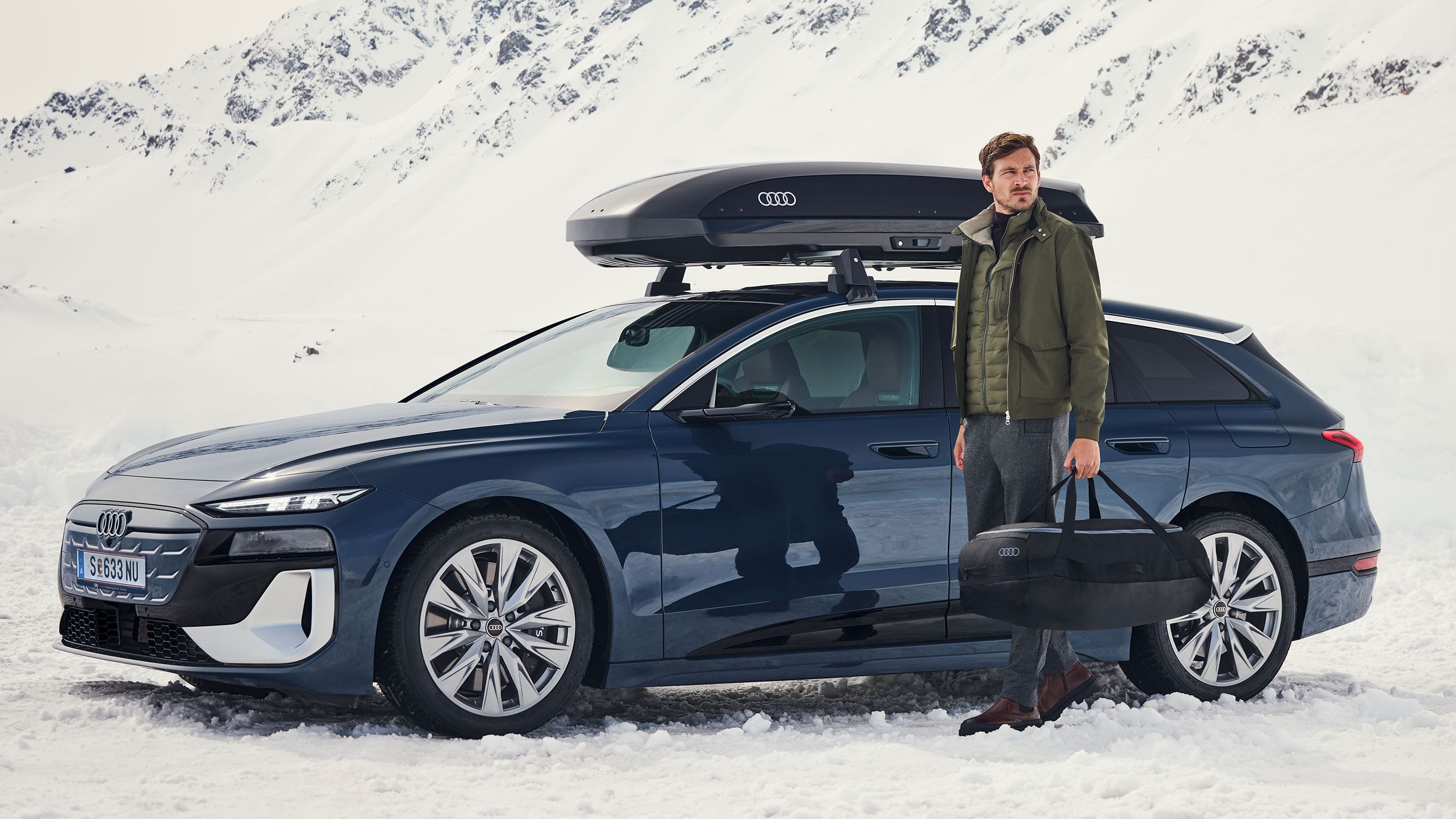 Ein Audi S6 mit Dachbox parkt in einer winterlichen Berglandschaft, ein Mann mit Tasche steht davor
