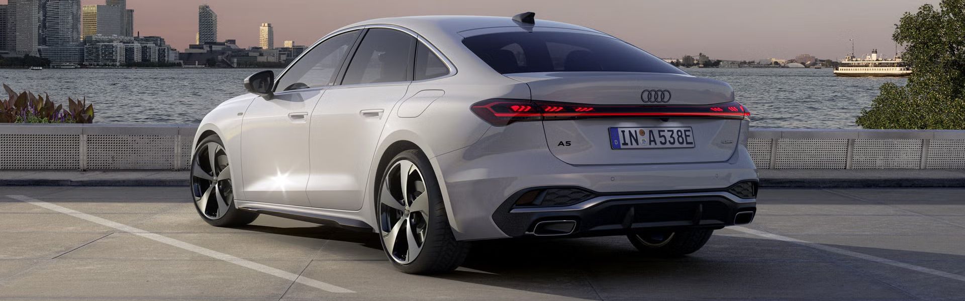 Die A5 Limousine e-hybrid steht im schrägen Heckprofil auf einem Parkplatz am Ufer eines Sees. Dahinter ist die Skyline einer Stadt bei Dämmerung zu sehen.