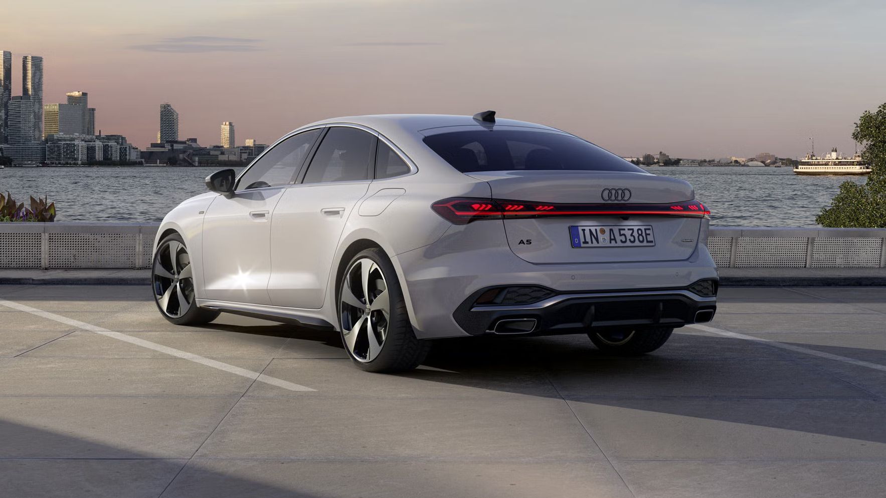 Die A5 Limousine e-hybrid steht im schrägen Heckprofil auf einem Parkplatz am Ufer eines Sees. Dahinter ist die Skyline einer Stadt bei Dämmerung zu sehen.