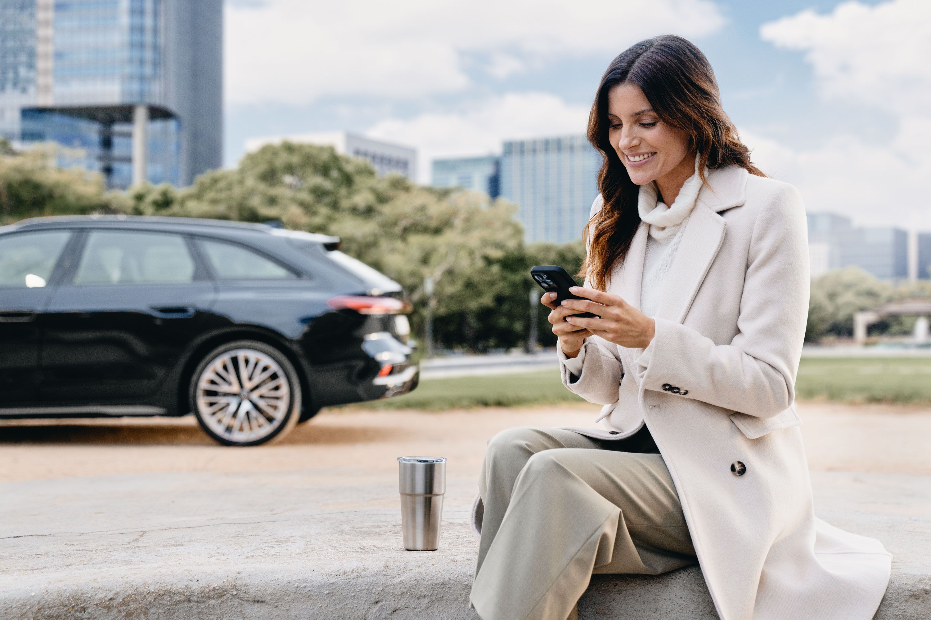 Eine Frau sitzt im Freien auf einer Steinmauer und schaut lächelnd auf ihr Smartphone. Im Hintergrund ist ein schwarzer Audi vor modernen Bürogebäuden und Bäumen geparkt.
