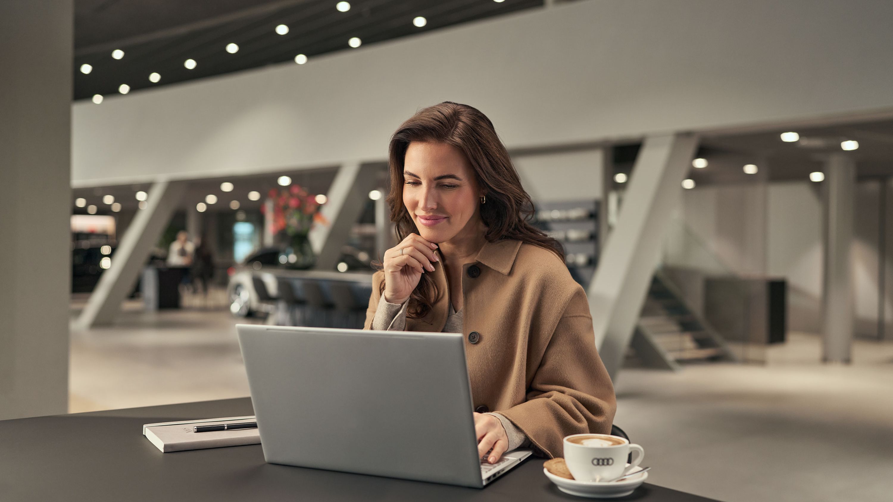Ein Kunde arbeitet in der Working Lounge an seinem Laptop und genießt dabei einen Kaffee; Notizbuch und Stift liegen auf dem Tisch.