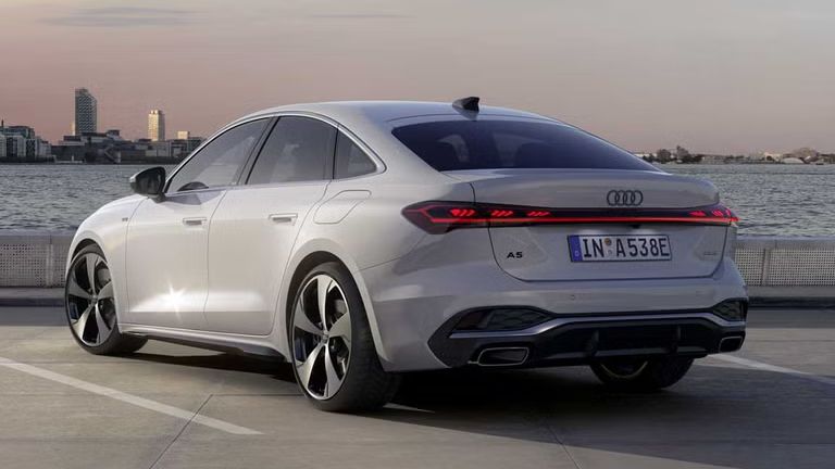 Die A5 Limousine e-hybrid steht im schrägen Heckprofil auf einem Parkplatz am Ufer eines Sees. Dahinter ist die Skyline einer Stadt bei Dämmerung zu sehen.