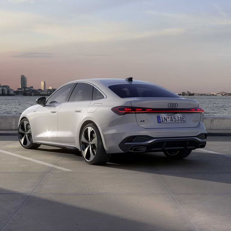 Die A5 Limousine e-hybrid steht im schrägen Heckprofil auf einem Parkplatz am Ufer eines Sees. Dahinter ist die Skyline einer Stadt bei Dämmerung zu sehen.