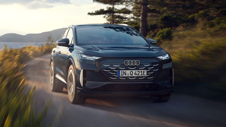 Der Q4 SUV e-tron schräg von vorn in voller Fahrt auf einer Landstraße an einem Waldsee, im Hintergrund eine bergige Landschaft.