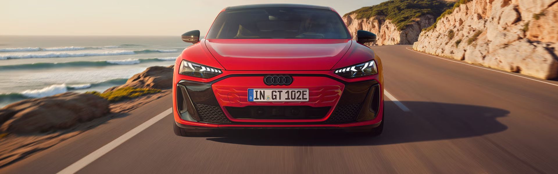 Frontansicht eines roten Audi e-tron GT quattro, der auf einer Küstenstraße fährt. Im Hintergrund sind Klippen, das Meer und der Himmel zu erkennen. 