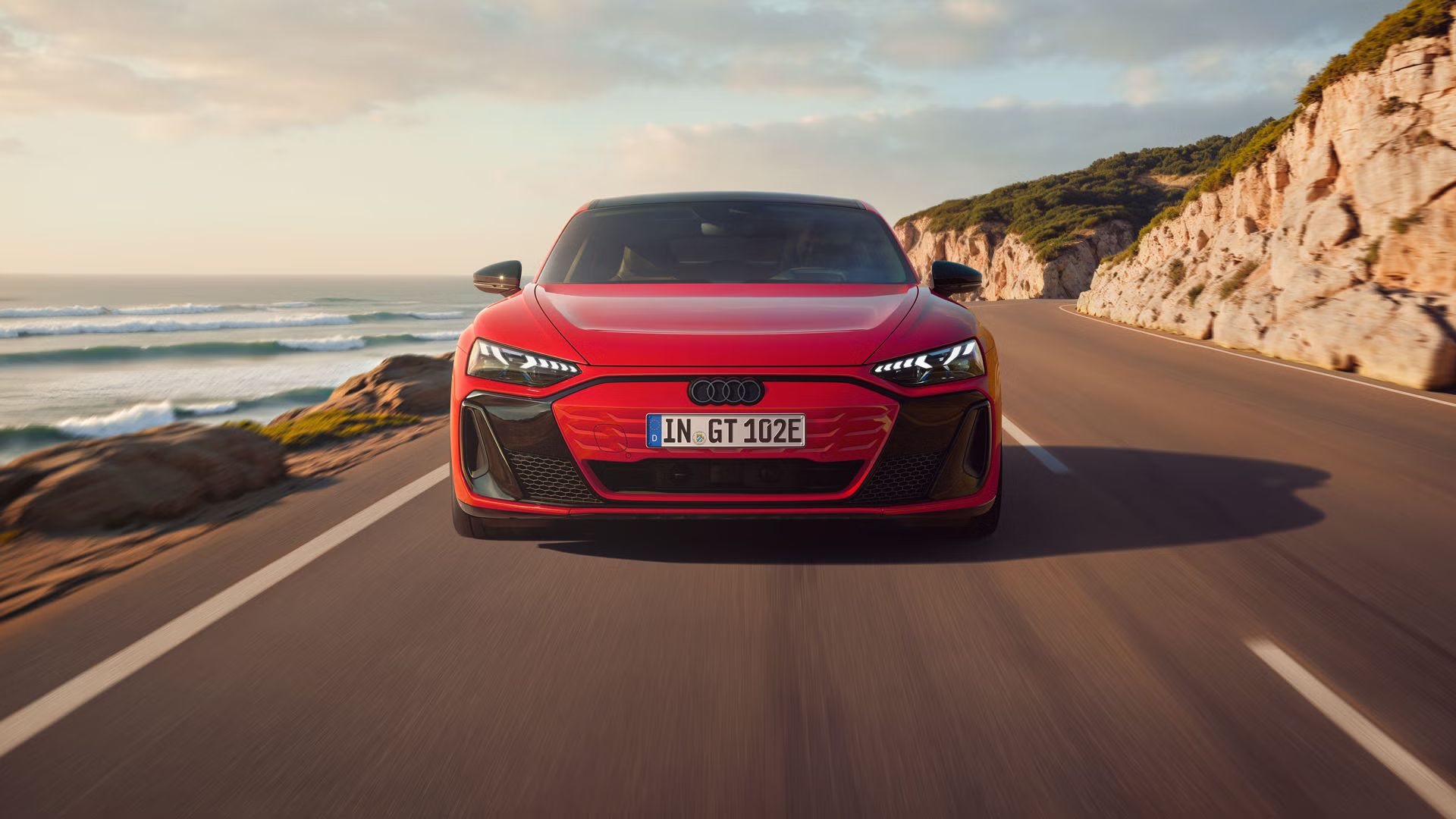 Frontansicht eines roten Audi e-tron GT quattro, der auf einer Küstenstraße fährt. Im Hintergrund sind Klippen, das Meer und der Himmel zu erkennen. 