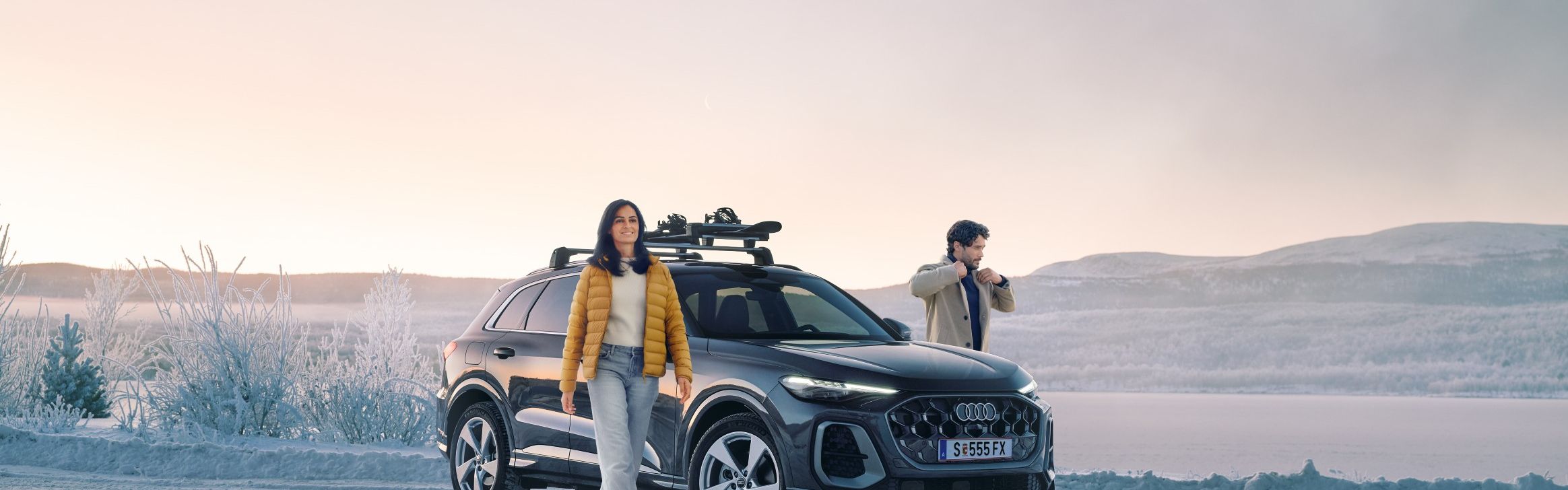Zwei Personen stehen auf einer verschneiten Straße neben einem schwarzen Audi Q5 mit Dachgepäckträger, im Hintergrund eine winterliche Landschaft mit zugefrorenem See und Hügeln. 