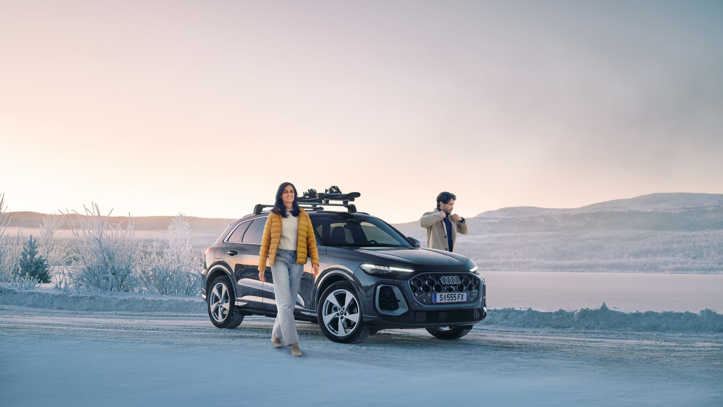 Zwei Personen stehen auf einer verschneiten Straße neben einem schwarzen Audi Q5 mit Dachgepäckträger, im Hintergrund eine winterliche Landschaft mit zugefrorenem See und Hügeln. 