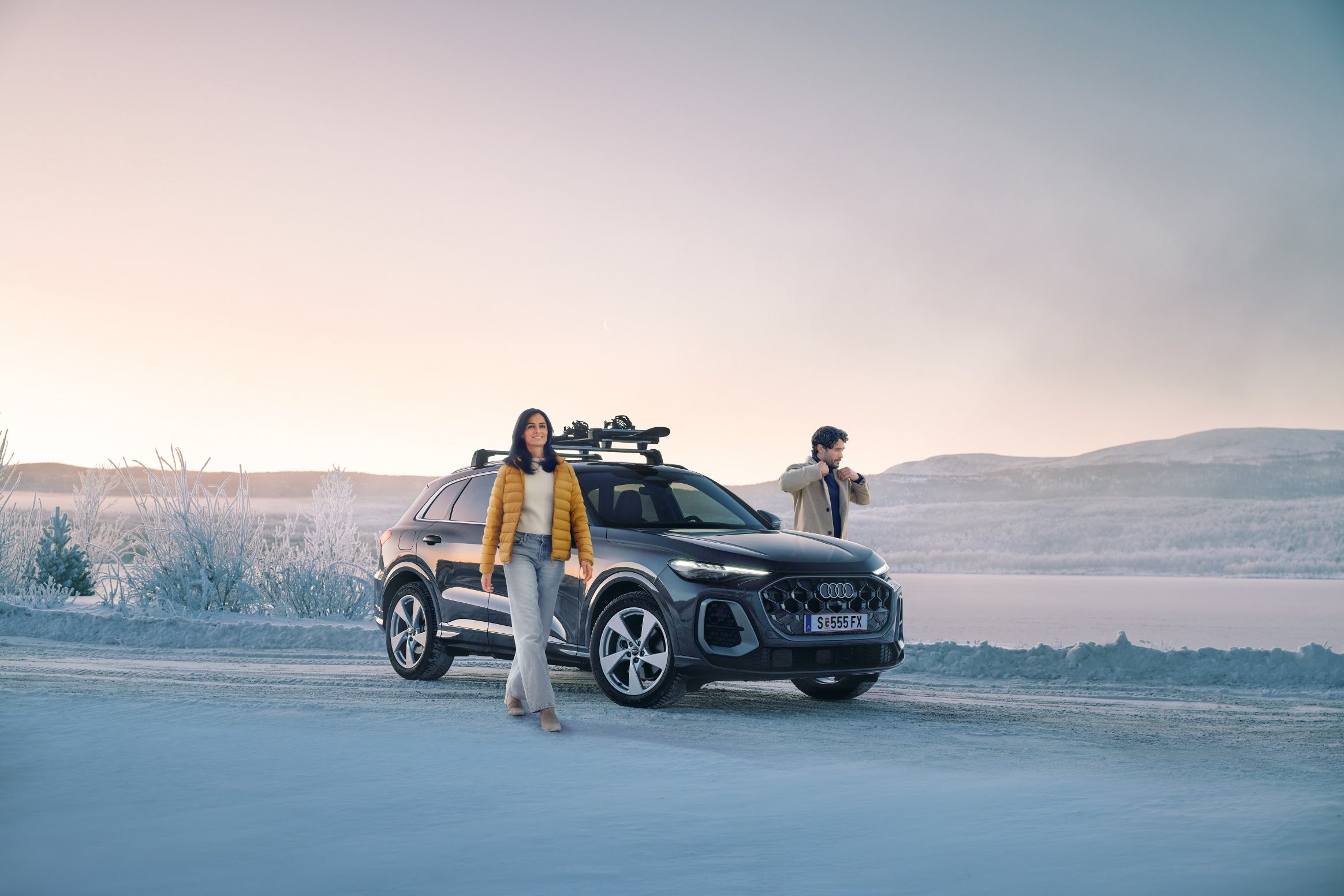 Zwei Personen stehen auf einer verschneiten Straße neben einem schwarzen Audi Q5 mit Dachgepäckträger, im Hintergrund eine winterliche Landschaft mit zugefrorenem See und Hügeln. 