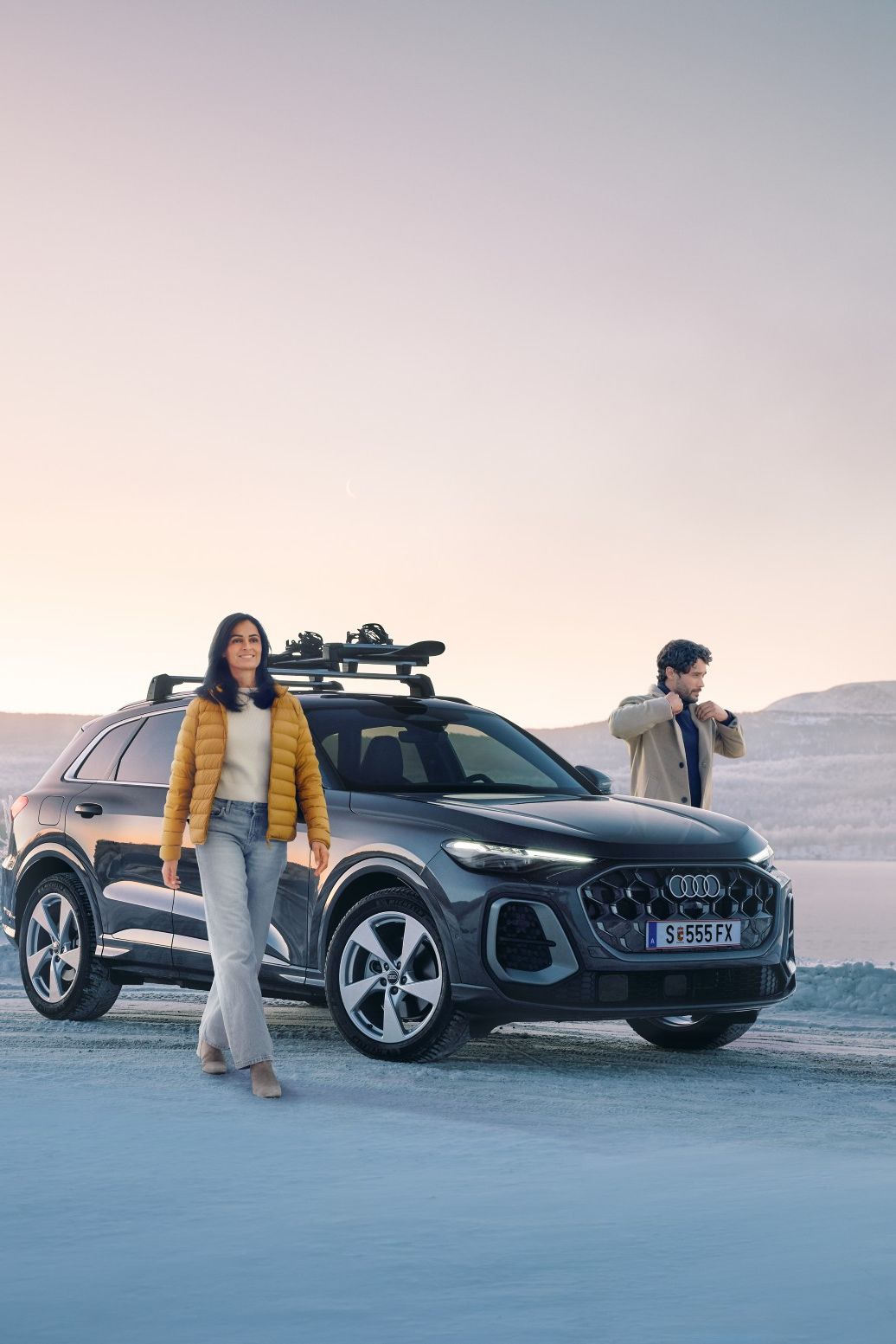 Zwei Personen stehen auf einer verschneiten Straße neben einem schwarzen Audi Q5 mit Dachgepäckträger, im Hintergrund eine winterliche Landschaft mit zugefrorenem See und Hügeln. 