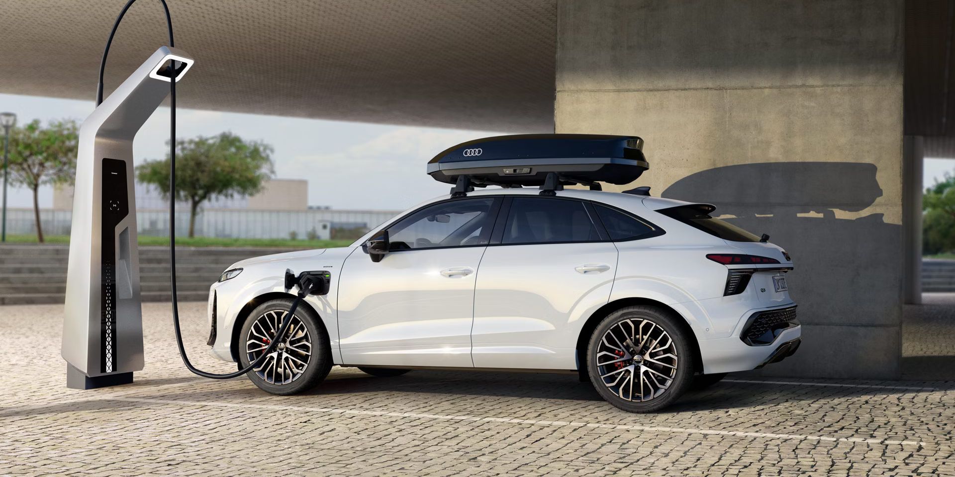Der Audi Q3 SUV Sportback von der Seite auf einem Parkplatz beim Laden. Weiterhin ist eine Dachbox auf dem weißen Auto zu erkennen.
