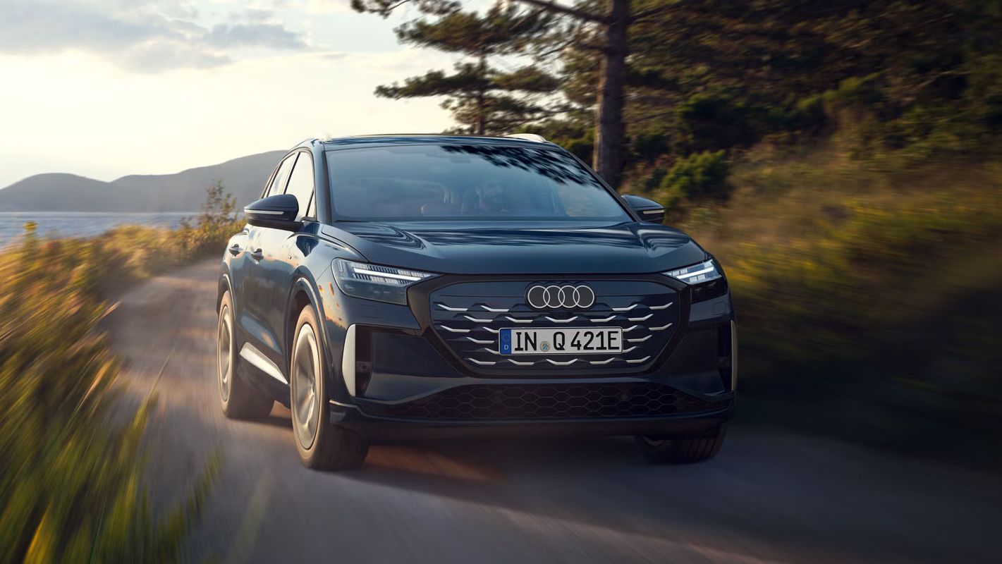 Der Q4 SUV e-tron schräg von vorn in voller Fahrt auf einer Landstraße an einem Waldsee, im Hintergrund eine bergige Landschaft.