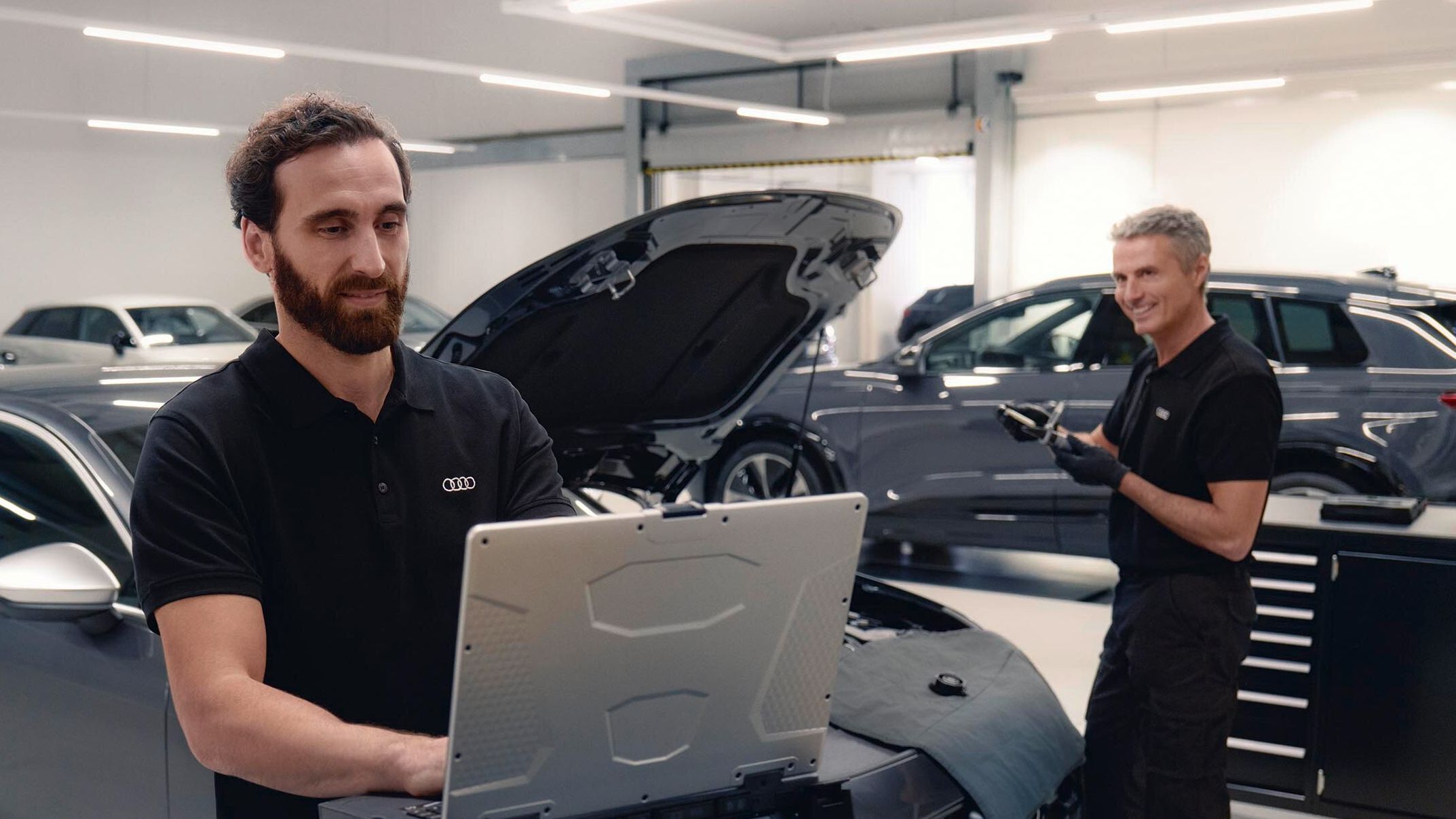 Zwei Audi Service Techniker in einer Werkstatt unterhalten sich. Einer von beiden blickt dabei auf seinen Laptop.