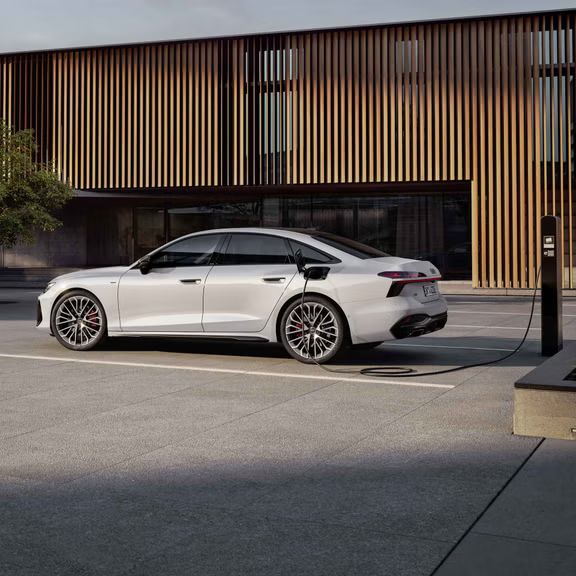 Die A6 Limousine e-hybrid in weiß steht seitlich vor einer Gebäudefassade und ist dabei an eine Ladestation angeschlossen.