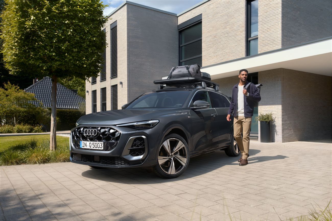 Ein Mann geht an einem in der Einfahrt eines Hauses parkenden Audi Q5 mit Dachkorb und darauf befestigten Audi Taschen vorbei.