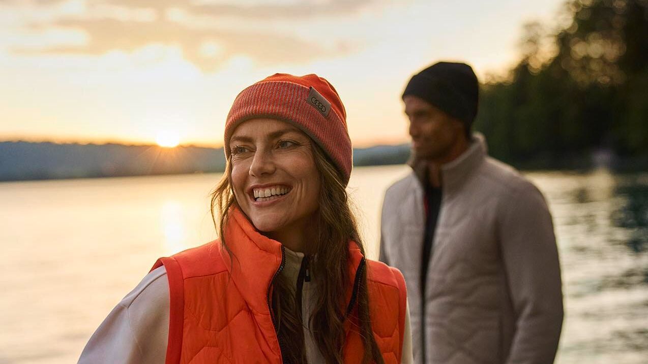 Eine Frau mit orangefarbener Steppweste und Mütze der Audi active collection steht lächelnd am Ufer eines Sees bei Sonnenuntergang. Hinter ihr steht ein Mann in heller Steppjacke und dunkler Mütze der Audi active collection, der auf das Wasser blickt. Die Stimmung ist ruhig und herbstlich.
