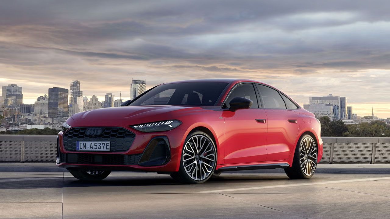 Die A5 Limousine e-hybrid im schrägen Frontprofil. Sie ist in misanorot lackiert. Im Hintergrund ist die Skyline einer Stadt bei Dämmerung zu sehen.