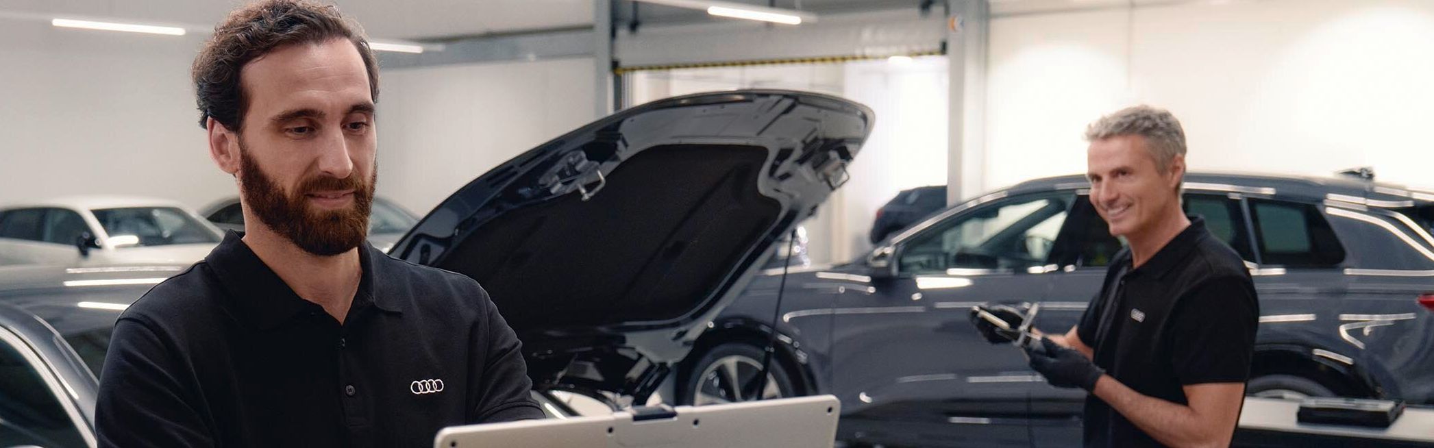 Zwei Audi Service Techniker in einer Werkstatt unterhalten sich. Einer von beiden blickt dabei auf seinen Laptop.