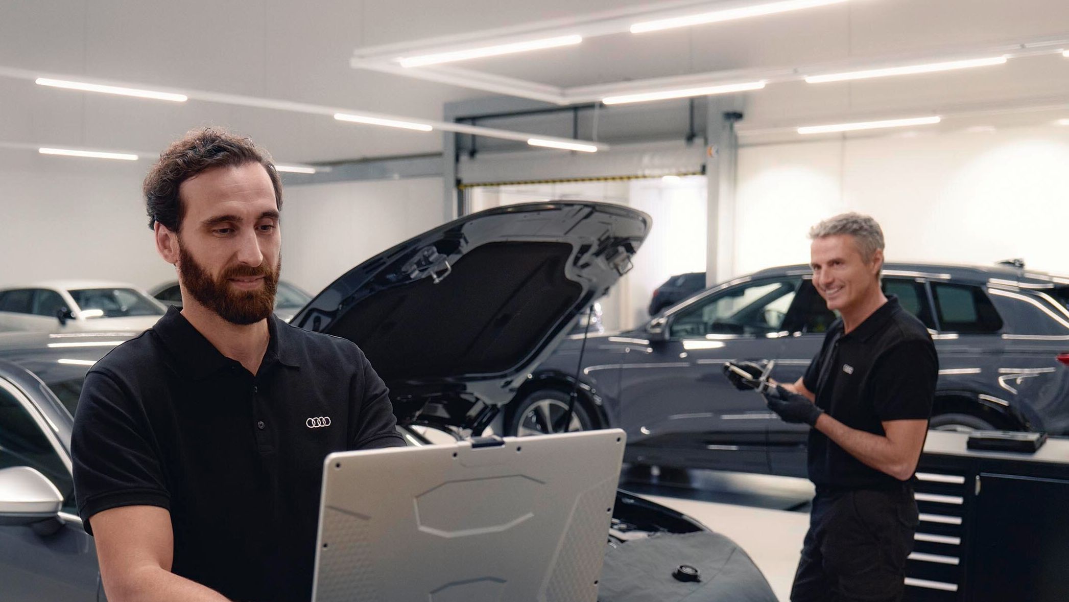 Zwei Audi Service Techniker in einer Werkstatt unterhalten sich. Einer von beiden blickt dabei auf seinen Laptop.