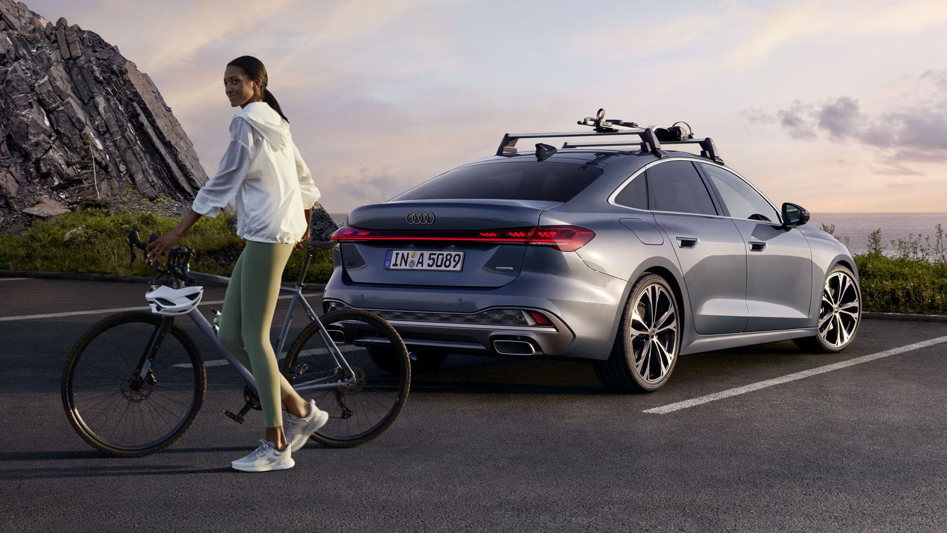 Die A5 Limousine im schrägen Heckprofil. Das Fahrzeug steht auf einem Parkplatz nahe eines Berges. Auf dem Dach befindet sich ein Fahrradhalter. Hinter dem Fahrzeug schiebt eine Junge Frau ein Fahrrad.