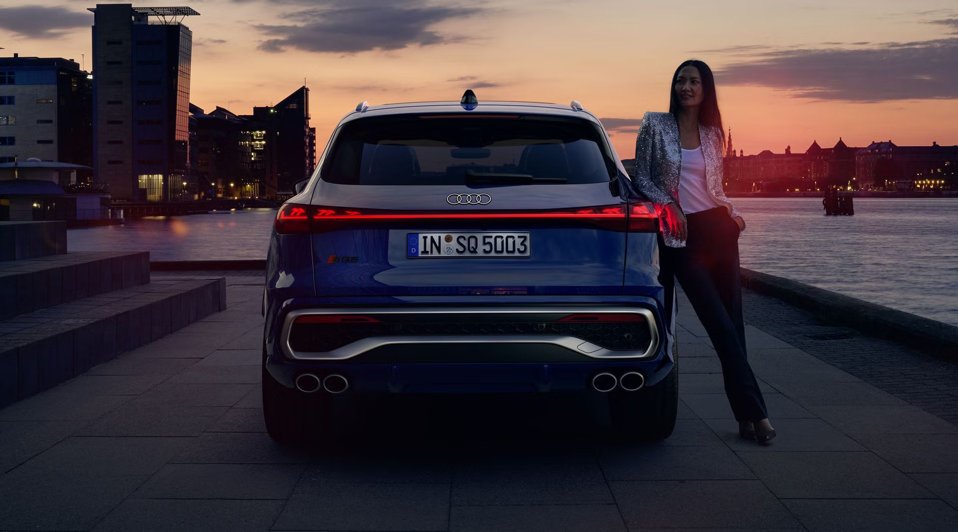 Der SQ5 SUV im Heckprofil vor der Skyline einer Küstenstadt. An das Heck gelehnt steht eine junge Frau.