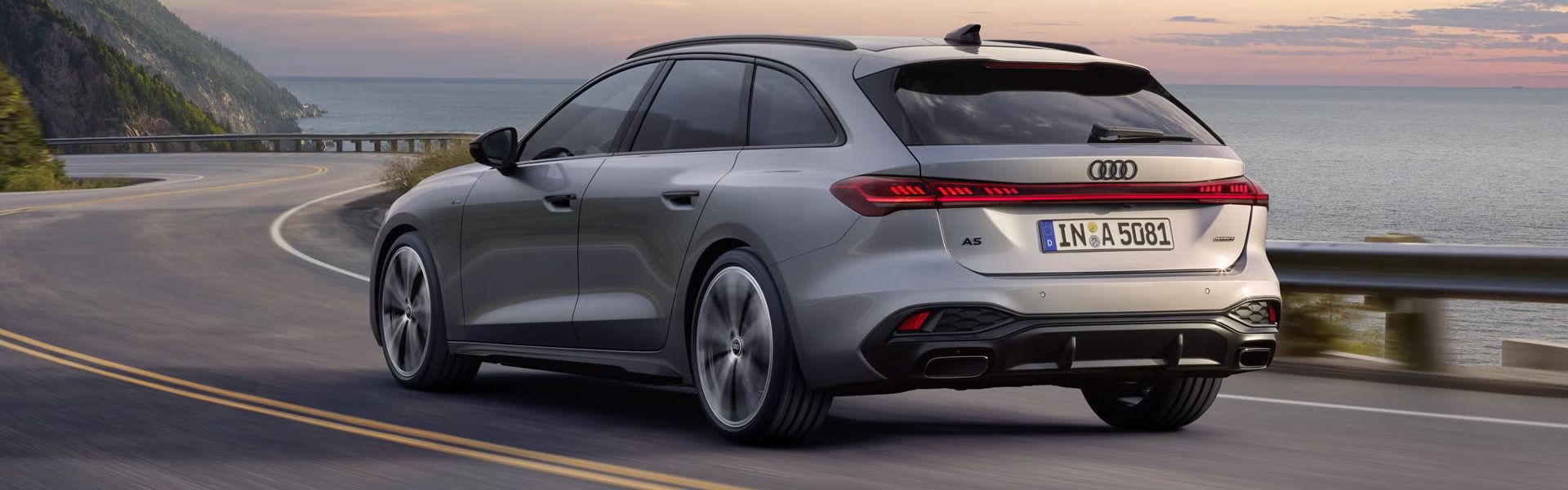Der A5 Avant in einer schrägen Heckperspektive, während er bei Sonnenuntergang eine kurvige Küstenstraße entlangfährt.