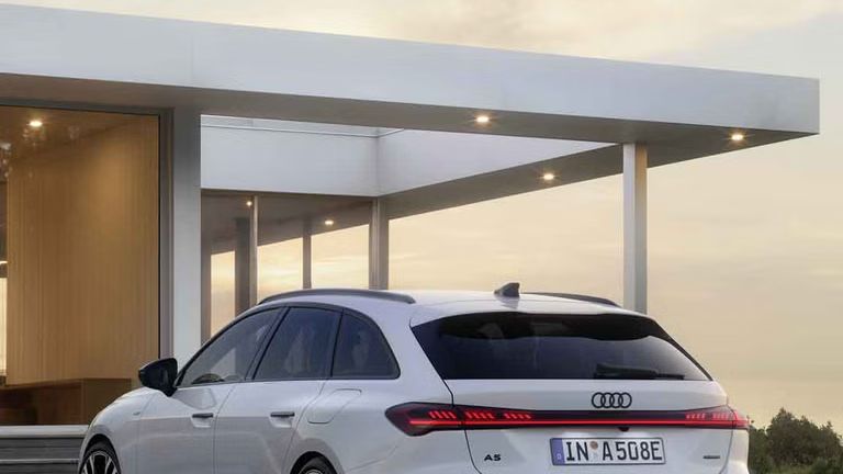 Der A5 Avant e-hybrid steht in einer schrägen Heckperspektive auf einer Kiesfläche vor einem modernen Wohnhaus.