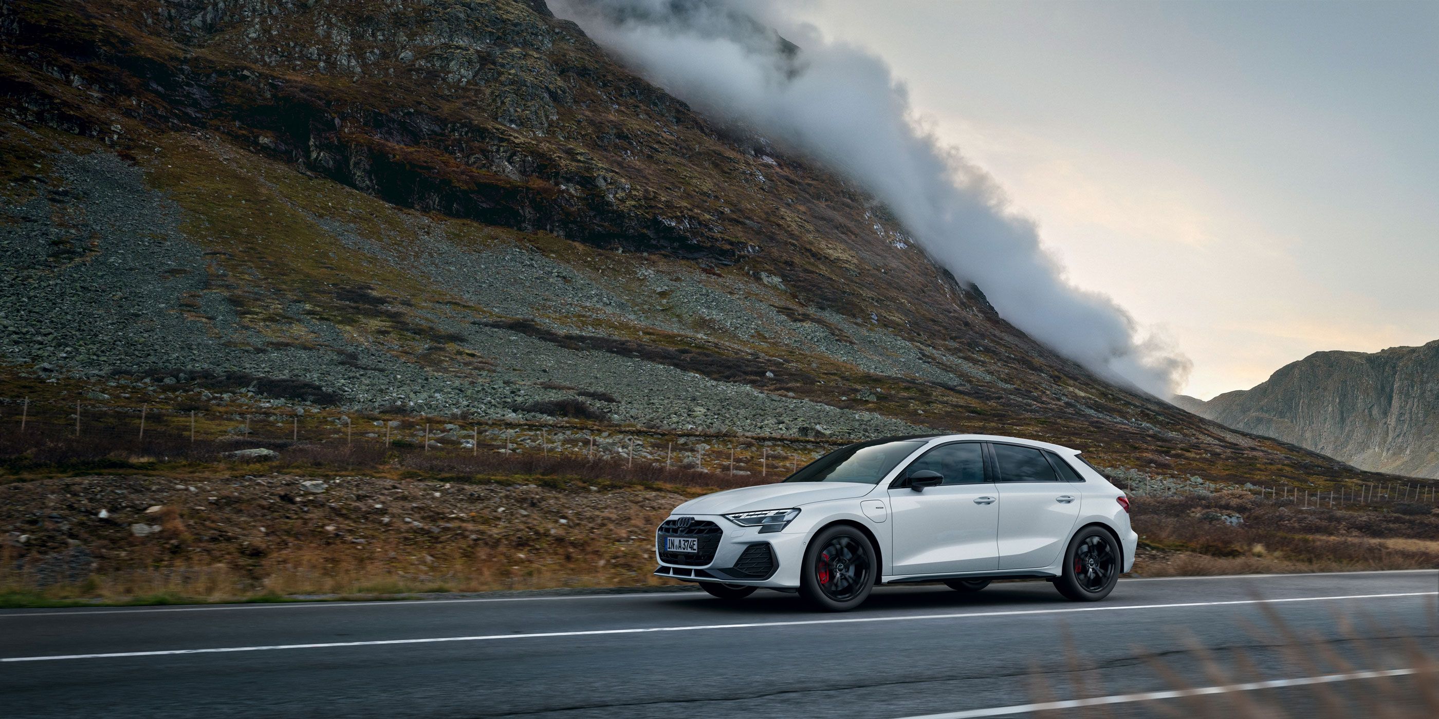Weißer Audi A3 Sportback e-hybrid fährt auf einer leeren Landstraße durch eine bergige Landschaft. Links erhebt sich ein steiler, felsiger Hang, aus dem sich Nebelschwaden lösen. Die Stimmung ist herbstlich und ruhig, mit gedämpften Farben und bewölktem Himmel.