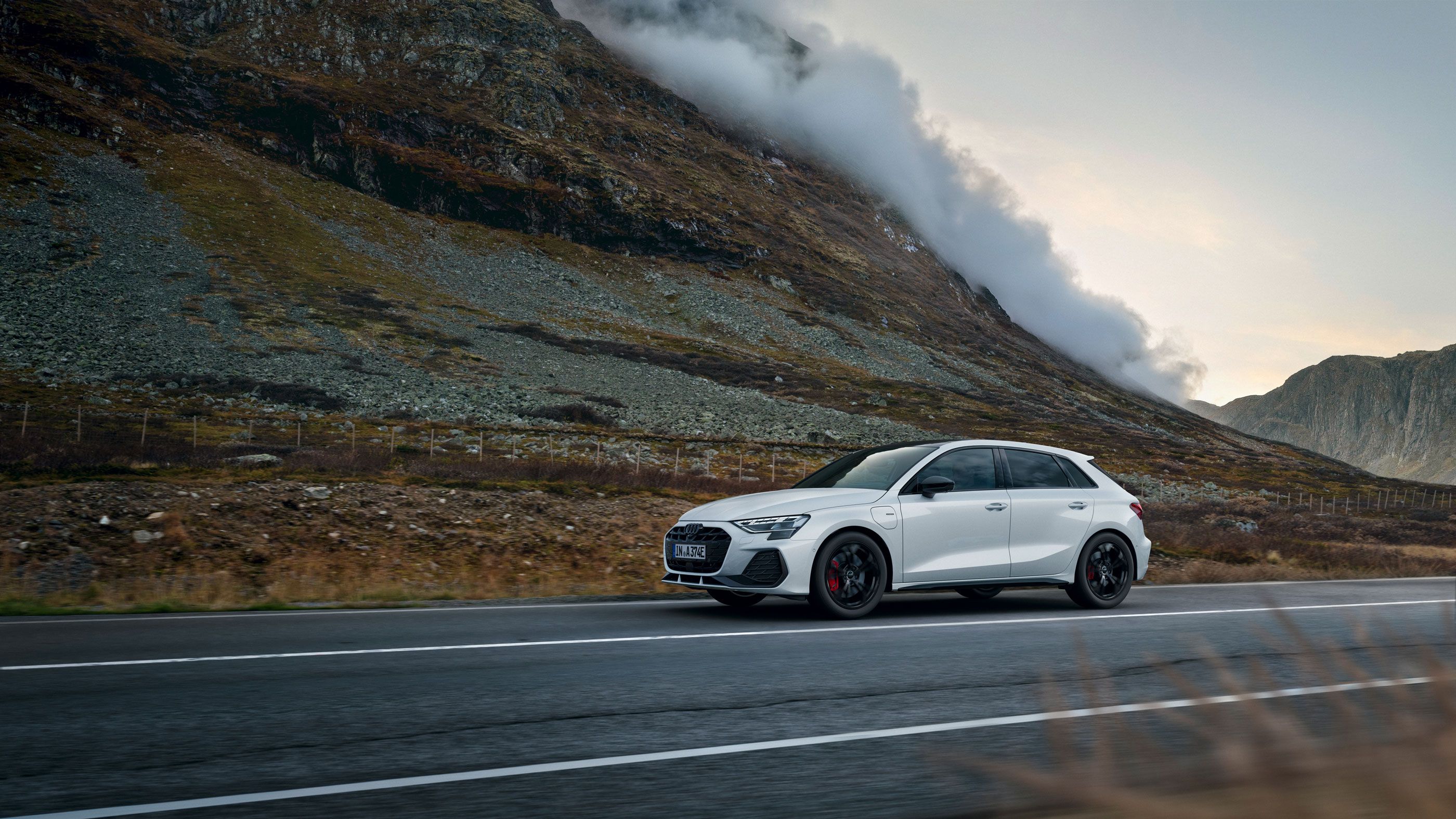 Weißer Audi A3 Sportback e-hybrid fährt auf einer leeren Landstraße durch eine bergige Landschaft. Links erhebt sich ein steiler, felsiger Hang, aus dem sich Nebelschwaden lösen. Die Stimmung ist herbstlich und ruhig, mit gedämpften Farben und bewölktem Himmel.