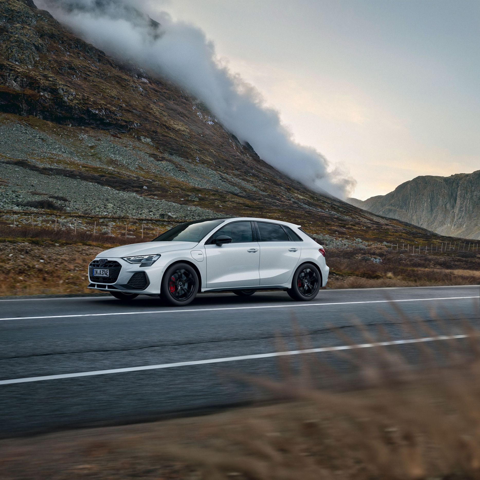 Weißer Audi A3 Sportback e-hybrid fährt auf einer leeren Landstraße durch eine bergige Landschaft. Links erhebt sich ein steiler, felsiger Hang, aus dem sich Nebelschwaden lösen. Die Stimmung ist herbstlich und ruhig, mit gedämpften Farben und bewölktem Himmel.