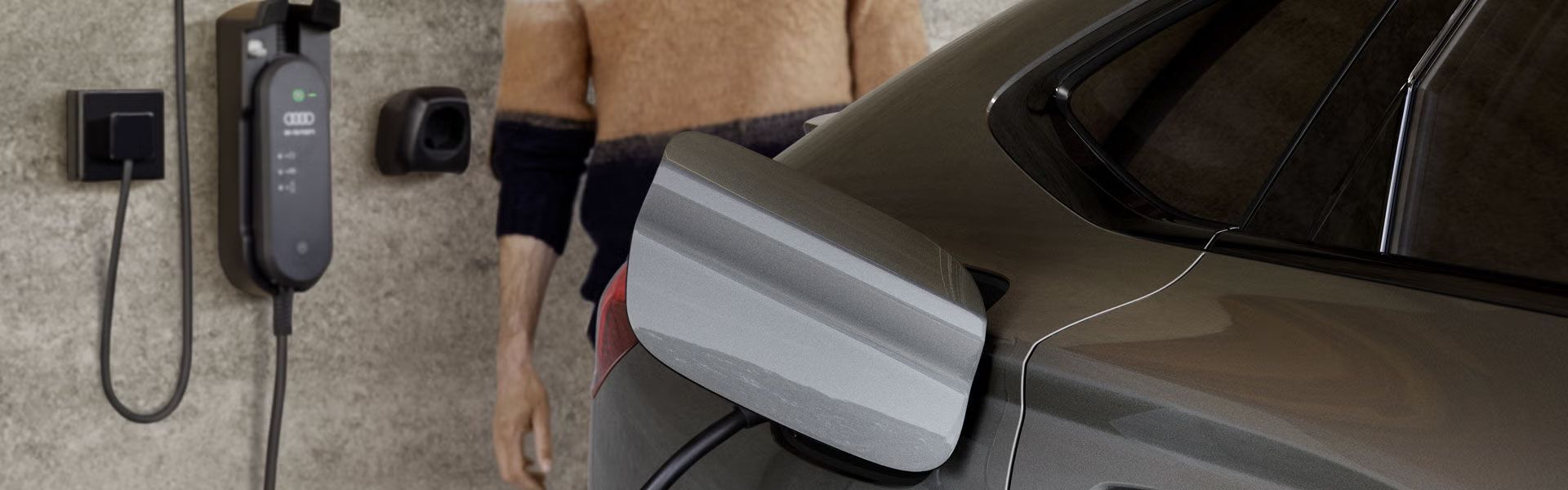 Nahaufnahme der Ladebuchse des Q6 Sportback e-tron, an welche eine an einer Hauswand montierte Ladestation angeschlossen ist. Neben der Ladestation ist ein Mann an die Hauswand gelehnt.