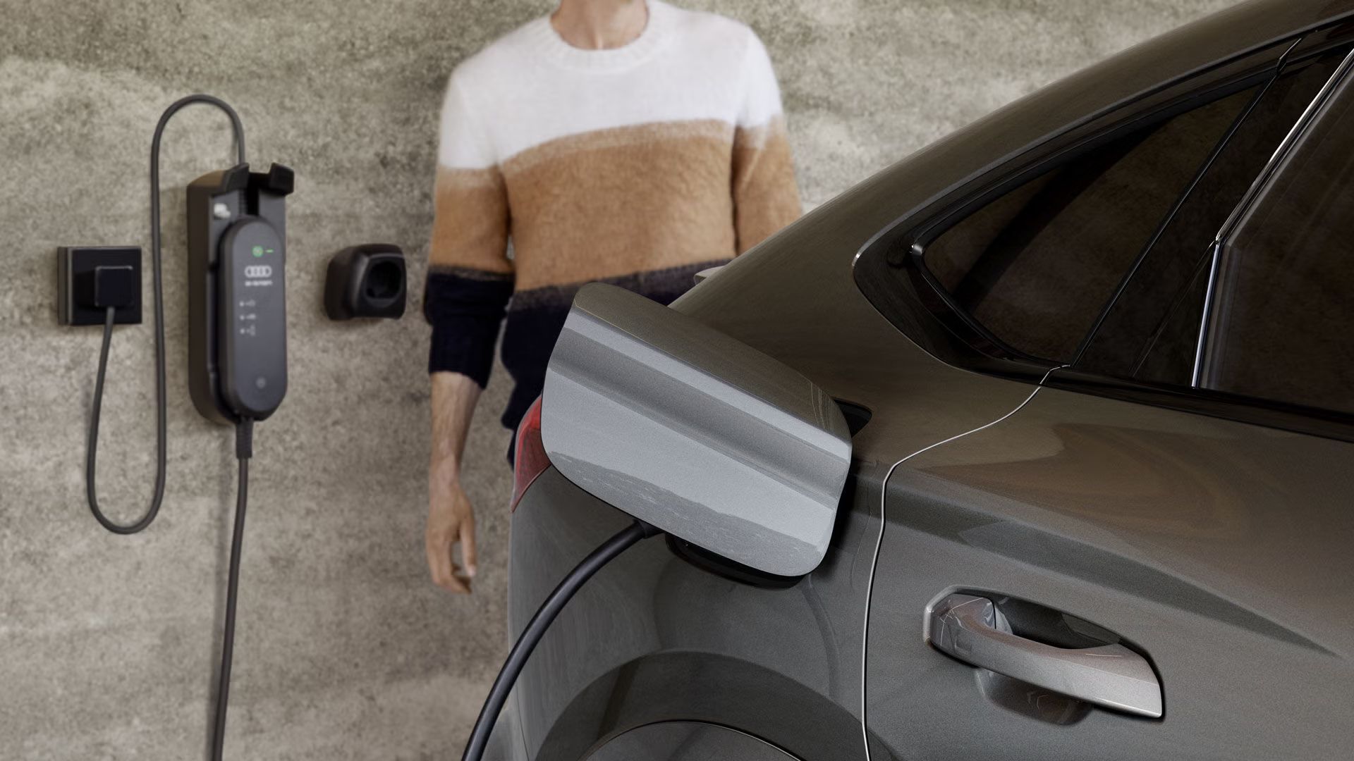 Nahaufnahme der Ladebuchse des Q6 Sportback e-tron, an welche eine an einer Hauswand montierte Ladestation angeschlossen ist. Neben der Ladestation ist ein Mann an die Hauswand gelehnt.